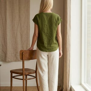 May include: An olive green short-sleeved top with a round neckline and a curved hemline paired with loose-fitting, full-length off-white trousers. The ensemble is shown from the back, highlighting the simple cut and the natural drape of the fabric.