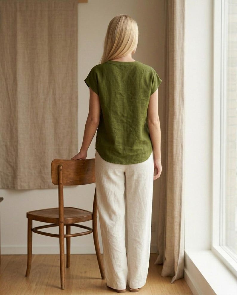 May include: An olive green short-sleeved top with a round neckline and a curved hemline paired with loose-fitting, full-length off-white trousers. The ensemble is shown from the back, highlighting the simple cut and the natural drape of the fabric.