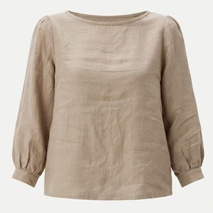 May include: A long-sleeved beige blouse featuring a round neckline, gathered shoulders, and buttoned cuffs. The garment is constructed from a textured fabric with a visible weave, showcasing a straight hemline and a relaxed, loose-fitting silhouette.