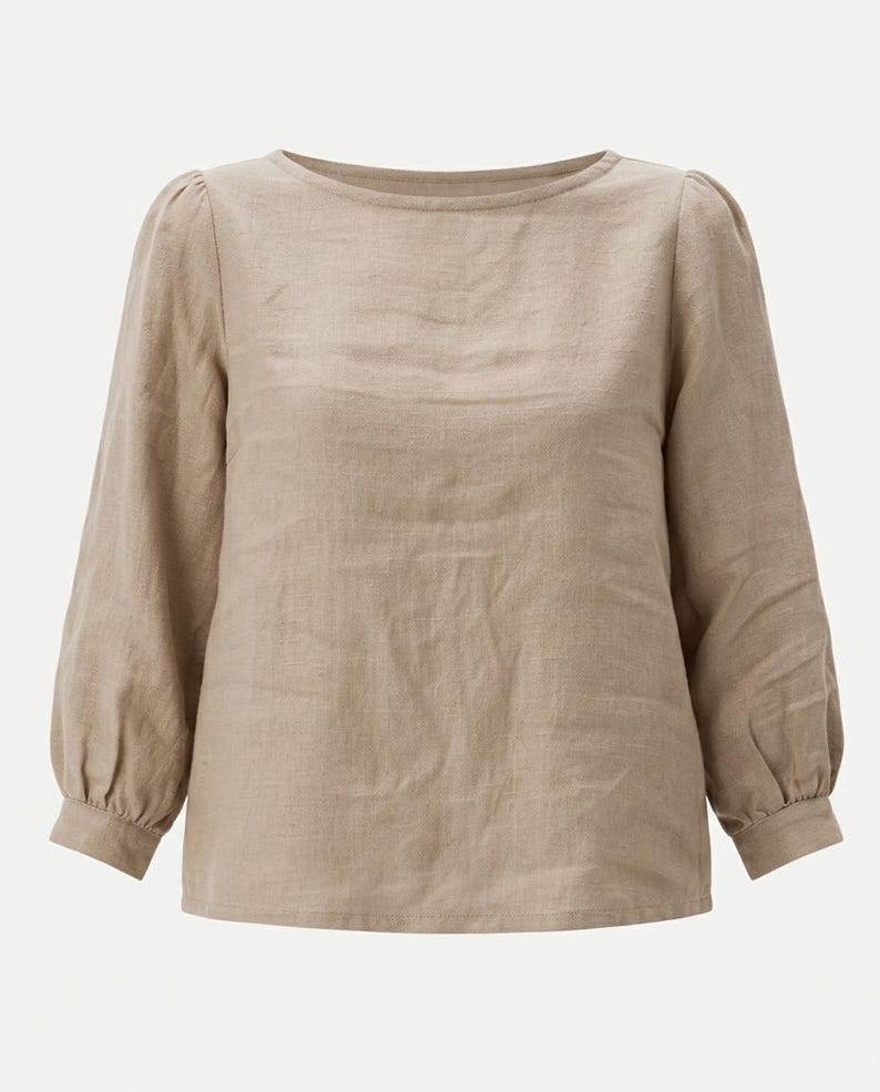 May include: A long-sleeved beige blouse featuring a round neckline, gathered shoulders, and buttoned cuffs. The garment is constructed from a textured fabric with a visible weave, showcasing a straight hemline and a relaxed, loose-fitting silhouette.