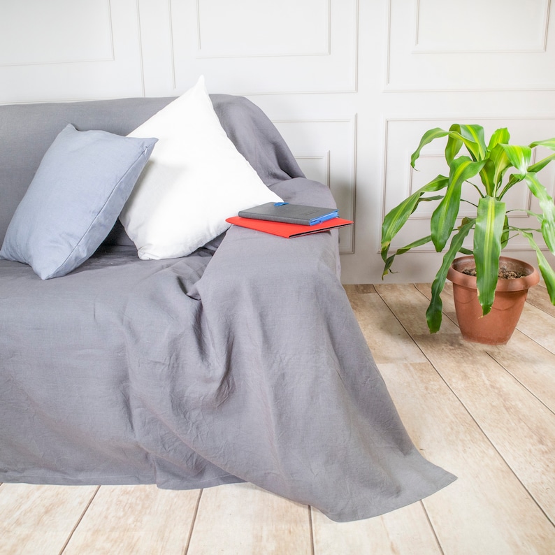 Puede incluir: Un cubre sof&aacute; de lino gris con dos almohadas, una blanca y una gris, y un cuaderno rojo y azul en la parte superior. El sof&aacute; est&aacute; frente a una pared blanca con lambrines. Una planta verde en una maceta de terracota est&aacute; en el suelo a la derecha del sof&aacute;.