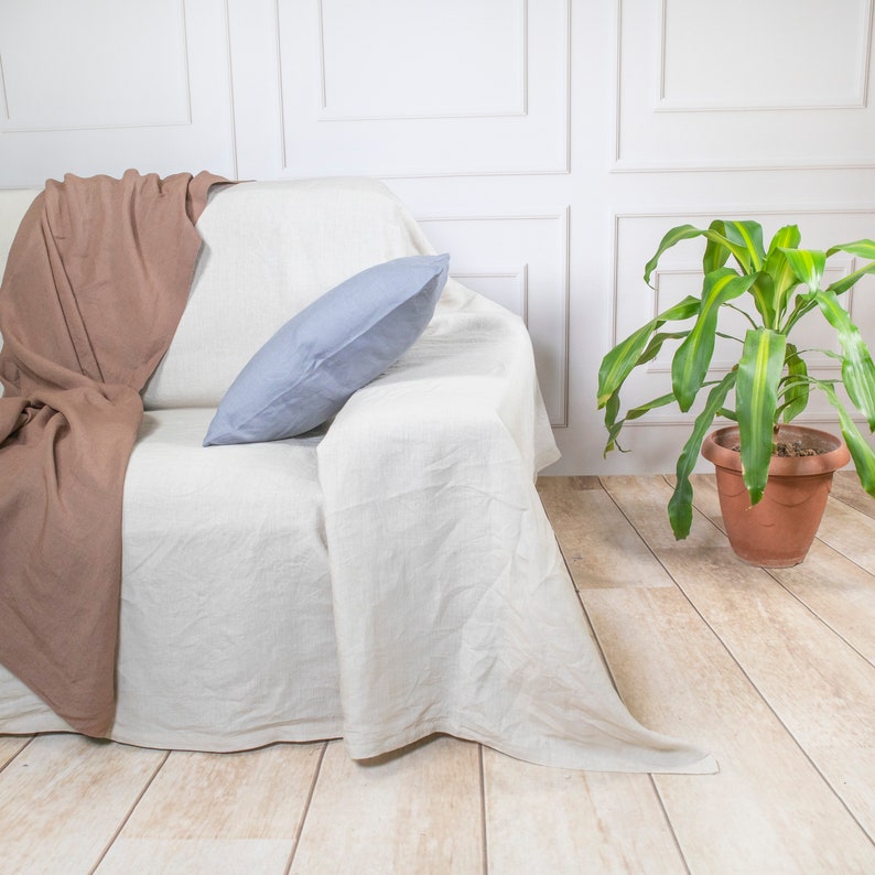 Puede incluir: Un sof&aacute; de lino blanco con una manta de tiro marr&oacute;n y una almohada azul claro. El sof&aacute; est&aacute; en una habitaci&oacute;n con paredes blancas y una planta en maceta.