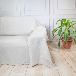 Puede incluir: Un cubre sof&aacute; de lino blanco drapeado sobre un sof&aacute; blanco. El cubre sof&aacute; est&aacute; doblado sobre el respaldo del sof&aacute; y cuelga por los lados. El sof&aacute; est&aacute; en una habitaci&oacute;n con paredes blancas y una planta verde en una maceta de terracota.