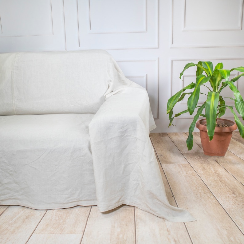 Puede incluir: Un cubre sof&aacute; de lino blanco drapeado sobre un sof&aacute; blanco. El cubre sof&aacute; est&aacute; doblado sobre el respaldo del sof&aacute; y cuelga por los lados. El sof&aacute; est&aacute; en una habitaci&oacute;n con paredes blancas y una planta verde en una maceta de terracota.