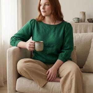 May include: A long-sleeved emerald green linen blouse with a round neckline and gathered cuffs, paired with loose-fitting tan linen trousers. The blouse features a simple, solid-colored design with a relaxed fit, while the trousers have a wide-leg cut.