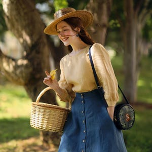 Może przedstawiać: Kobieta w słomkowym kapeluszu, jasnożółtej bluzie i niebieskiej dżinsowej spódnicy z guzikami z przodu. Trzyma wiklinowy kosz piknikowy i małą okrągłą torebkę z kwiatowym wzorem.