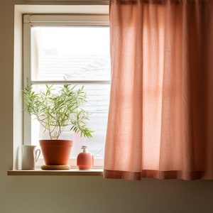 May include: A potted green plant sits on a windowsill with a white pitcher and a pink ceramic bottle. A peach-colored curtain hangs in the window behind the plant.