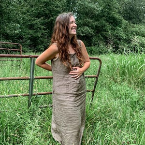 May include: A woman in a brown linen dress stands in a field of tall grass, smiling and looking to the right. She is standing near a rusty metal gate.