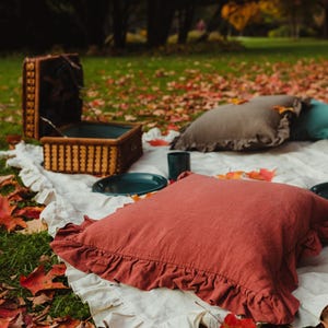 Puede incluir: Una escena de picnic r&uacute;stica con una manta blanca, dos almohadas, una cesta de mimbre y un plato azul verdoso sobre un lecho de hojas de oto&ntilde;o ca&iacute;das.