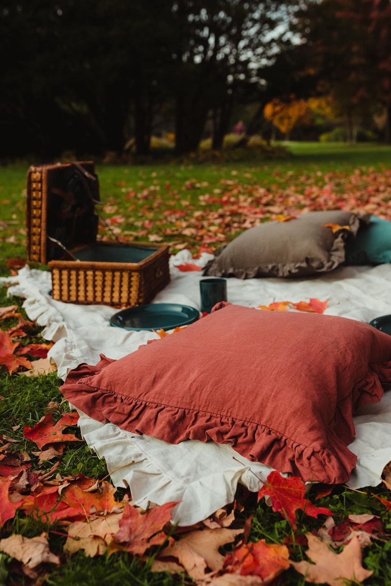 Puede incluir: Una escena de picnic r&uacute;stica con una manta blanca, dos almohadas, una cesta de mimbre y un plato azul verdoso sobre un lecho de hojas de oto&ntilde;o ca&iacute;das.