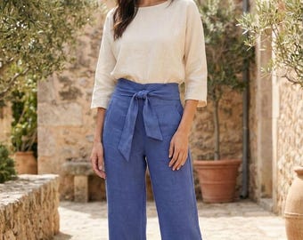 Puede incluir: Una mujer con una blusa de lino color crema con mangas tres cuartos y pantalones de lino azules. Los pantalones tienen cintura alta y cinturón de lazo a juego. El conjunto es de tejido ligero, perfecto para el clima cálido.