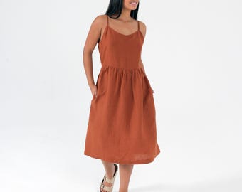 Puede incluir: Un vestido midi color óxido con tirantes finos y cintura fruncida. El vestido tiene bolsillos y llega hasta la pantorrilla. La persona lleva sandalias beige.