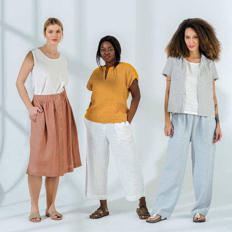 Pu&ograve; includere: Tre donne che indossano abiti di lino. La donna a sinistra indossa una canotta bianca senza maniche e una gonna marrone. La donna al centro indossa una blusa gialla e pantaloni bianchi. La donna a destra indossa una camicia bianca con una camicia a quadri sopra e pantaloni blu.