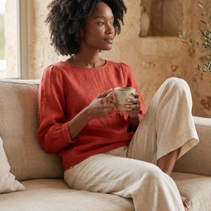 May include: A rust-colored long-sleeved blouse with a round neckline and gathered sleeve cuffs, paired with loose-fitting beige trousers. The blouse features a simple, solid-colored design with a relaxed fit, while the trousers are light-toned.