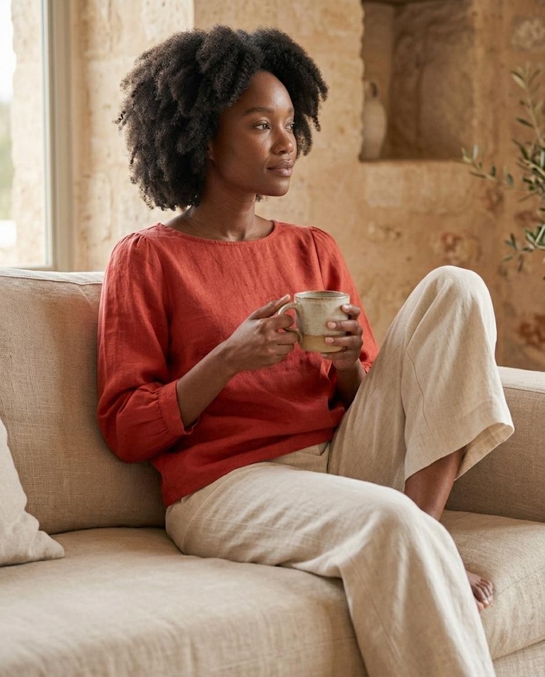 May include: A rust-colored long-sleeved blouse with a round neckline and gathered sleeve cuffs, paired with loose-fitting beige trousers. The blouse features a simple, solid-colored design with a relaxed fit, while the trousers are light-toned.
