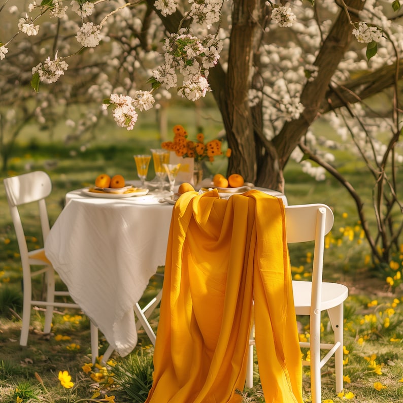 May include: A white tablecloth with a yellow cloth napkin on a table set for two. The table is set with two white plates, two glasses, and two oranges. The table is set in a garden with a blooming tree in the background.