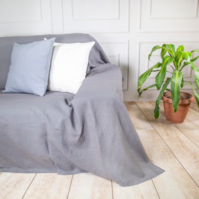 Puede incluir: Un cubre sof&aacute; de lino gris con dos almohadas, una blanca y una gris, sobre un suelo de baldosas blancas y grises. Una planta verde en una maceta de terracota est&aacute; en el fondo.