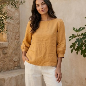 May include: A mustard yellow long-sleeved blouse with a round neckline and gathered cuffs paired with off-white trousers. The blouse features a straight hemline and a simple, solid-colored design, while the trousers have a clean, tailored fit.