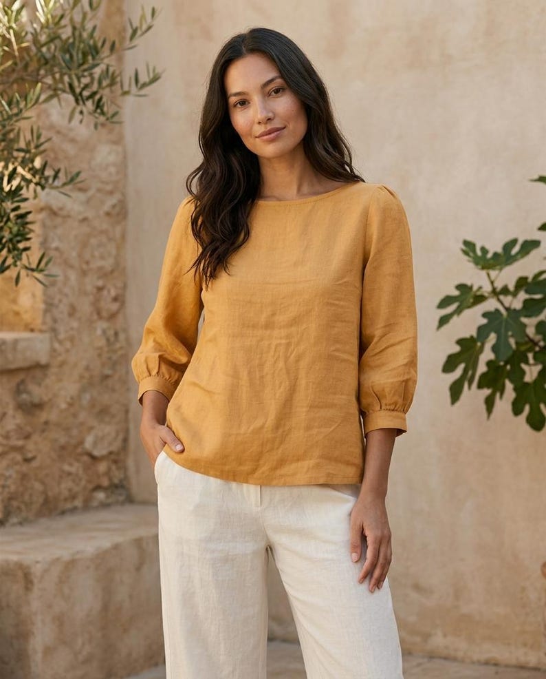 May include: A mustard yellow long-sleeved blouse with a round neckline and gathered cuffs paired with off-white trousers. The blouse features a straight hemline and a simple, solid-colored design, while the trousers have a clean, tailored fit.