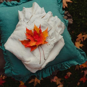 Puede incluir: Una almohada de lino blanco con un borde con volantes se coloca sobre una almohada turquesa con un borde con volantes. Un grupo de hojas de oto&ntilde;o naranja y rojas se coloca sobre la almohada blanca.
