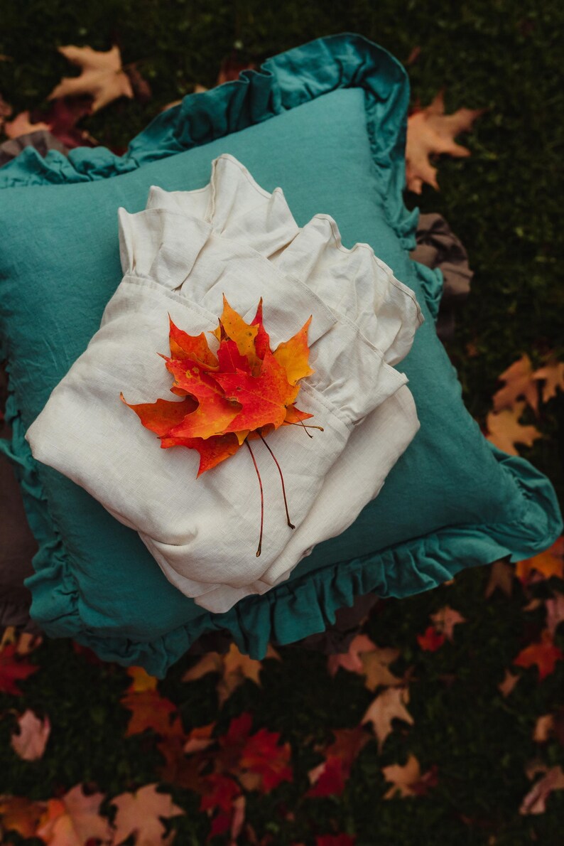 Puede incluir: Una almohada de lino blanco con un borde con volantes se coloca sobre una almohada turquesa con un borde con volantes. Un grupo de hojas de oto&ntilde;o naranja y rojas se coloca sobre la almohada blanca.