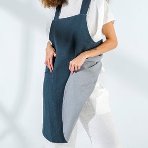 Pu&ograve; includere: Un grembiule in lino blu scuro con una grande tasca sul davanti e un pannello in lino grigio sul lato. Il grembiule &egrave; indossato sopra una camicia bianca.
