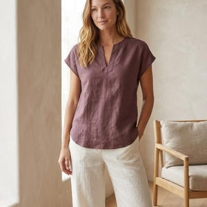 May include: A short-sleeved, plum-colored linen blouse featuring a notched V-neckline and a relaxed fit. The top has a straight hemline and is paired with loose-fitting, off-white linen trousers that feature a clean, simple design.