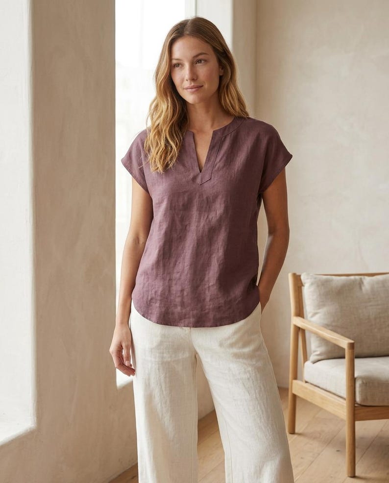 May include: A short-sleeved, plum-colored linen blouse featuring a notched V-neckline and a relaxed fit. The top has a straight hemline and is paired with loose-fitting, off-white linen trousers that feature a clean, simple design.
