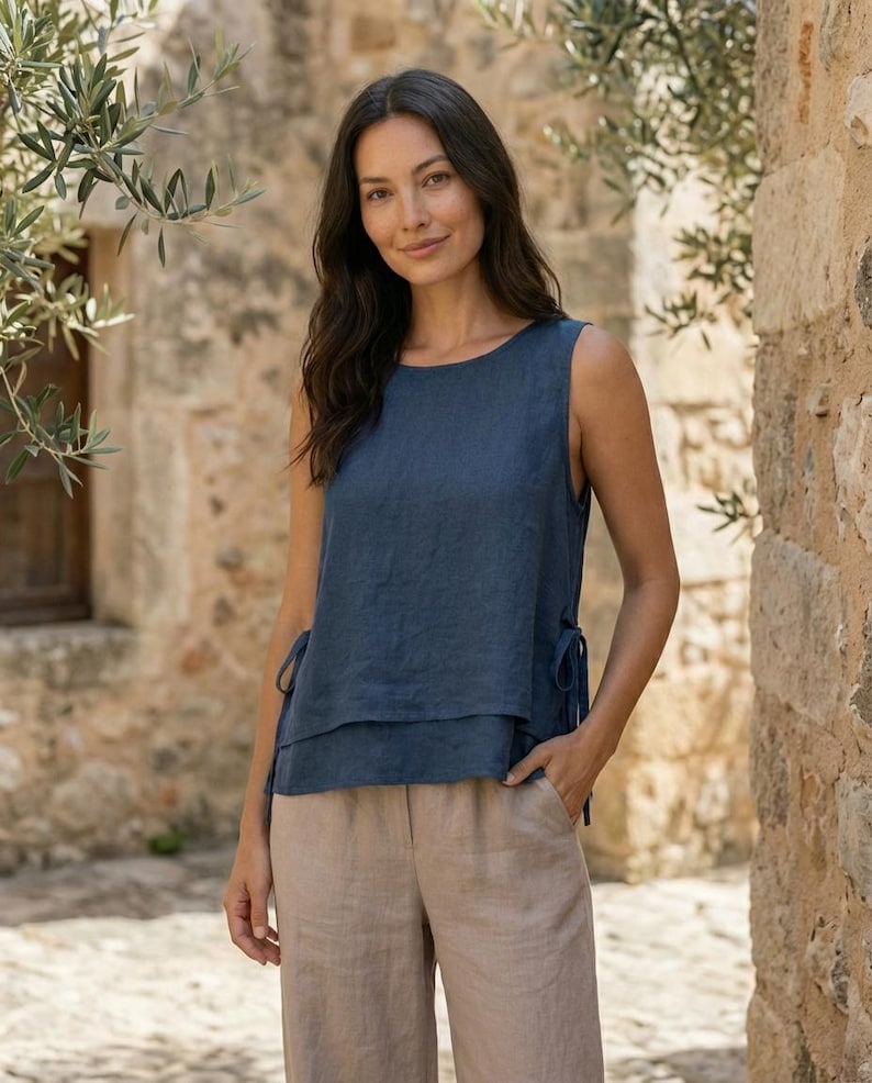 Puede incluir: Una blusa sin mangas de lino azul marino con un dise&ntilde;o en capas y lazos laterales. La modelo lleva la blusa con pantalones de lino beige. El fondo es una pared de piedra con ramas de olivo.
