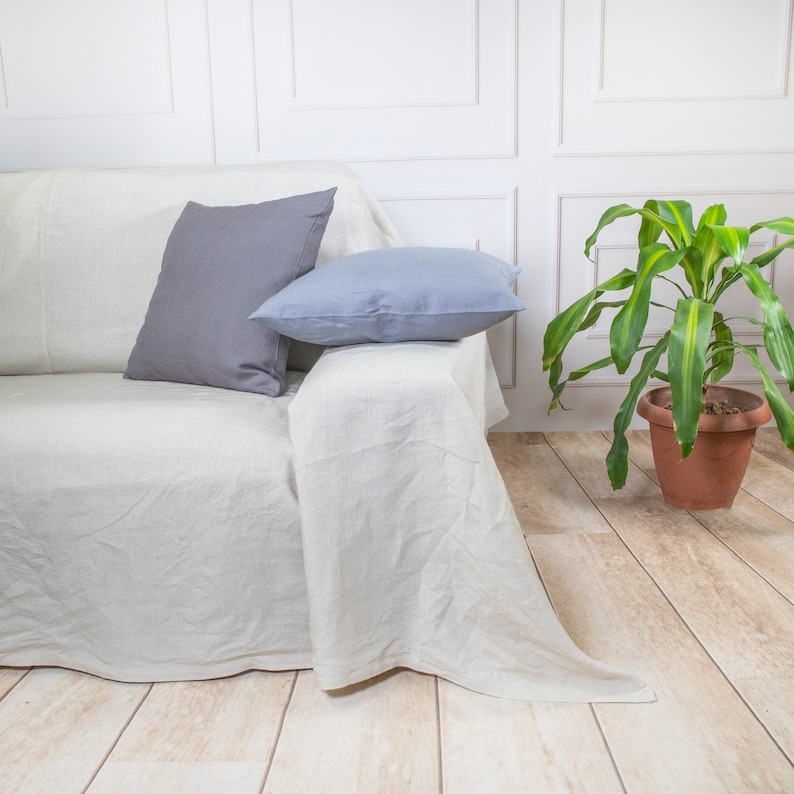 Puede incluir: Un sof&aacute; de lino blanco con dos cojines, uno gris y otro azul claro, contra una pared blanca con una planta verde en una maceta de terracota.
