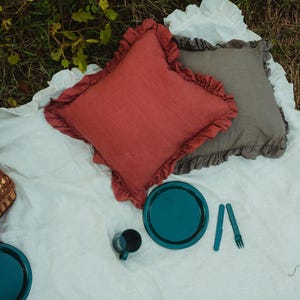Puede incluir: Un mantel de picnic blanco con tres almohadas, dos azul turquesa y una roja, con un plato azul turquesa y una taza encima.