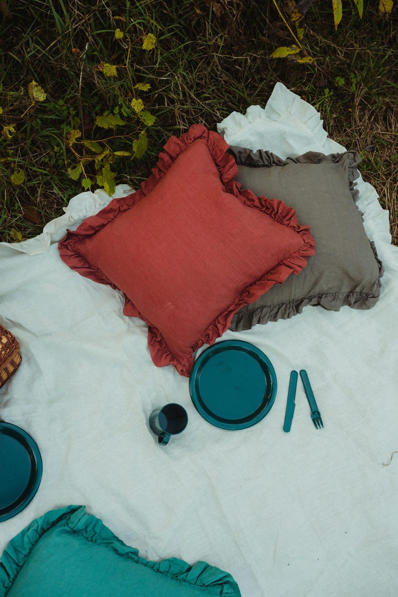 Puede incluir: Un mantel de picnic blanco con tres almohadas, dos azul turquesa y una roja, con un plato azul turquesa y una taza encima.