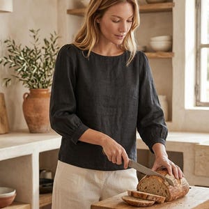 May include: A charcoal gray long-sleeved linen blouse with a round neckline and gathered sleeve cuffs, paired with light beige linen trousers. The garment features a simple, solid-colored design with a relaxed fit and visible fabric texture.