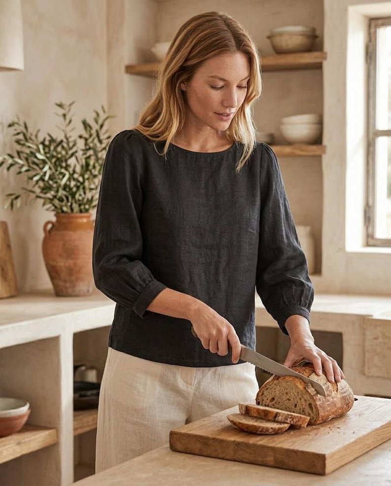 May include: A charcoal gray long-sleeved linen blouse with a round neckline and gathered sleeve cuffs, paired with light beige linen trousers. The garment features a simple, solid-colored design with a relaxed fit and visible fabric texture.