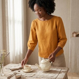 May include: A mustard yellow long-sleeved blouse with a round neckline and gathered sleeve cuffs, paired with light beige trousers. The blouse features a simple, solid-colored design with a relaxed fit and three-quarter length sleeves.