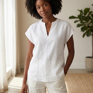 May include: A white short-sleeved blouse featuring a split V-neckline and a relaxed fit. The top is constructed from a textured fabric with a straight hemline that falls at the hip, providing a clean and simple silhouette for everyday wear.