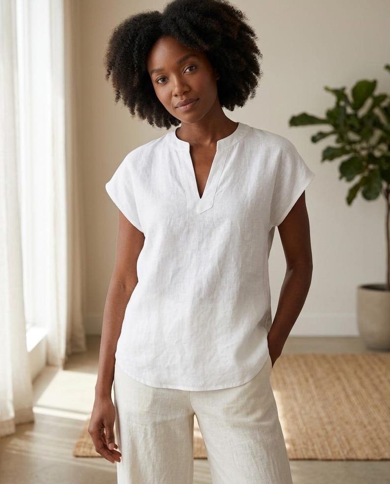 May include: A white short-sleeved blouse featuring a split V-neckline and a relaxed fit. The top is constructed from a textured fabric with a straight hemline that falls at the hip, providing a clean and simple silhouette for everyday wear.