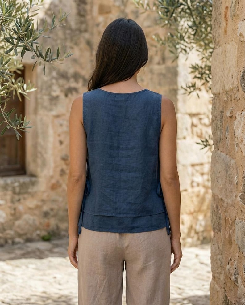 Puede incluir: Una blusa sin mangas de lino azul marino con un dise&ntilde;o en capas. Se combina con pantalones de lino color beige. La modelo est&aacute; de pie frente a una pared de piedra y ramas de olivo.