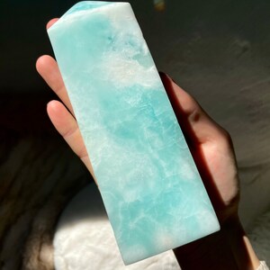 May include: A polished, tower-shaped crystal with a smooth, rectangular base and a pointed top. The stone features a vibrant, translucent light blue color with marbled white inclusions and a natural, crystalline texture throughout its surface.