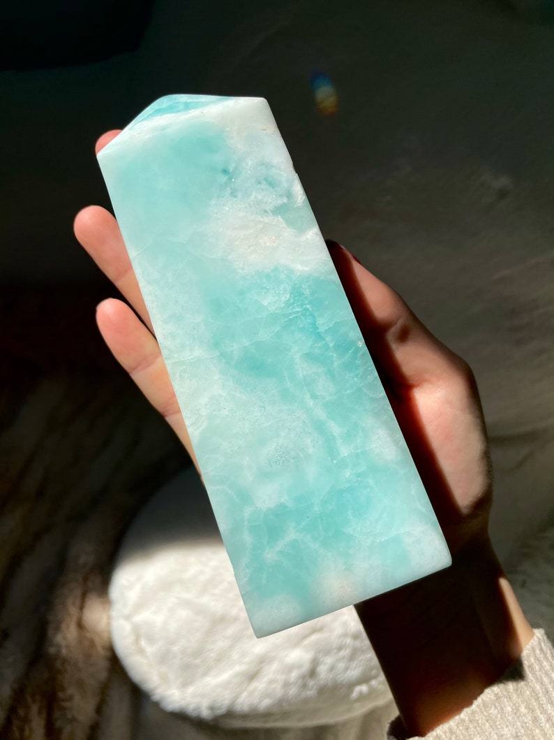 May include: A polished, tower-shaped crystal with a smooth, rectangular base and a pointed top. The stone features a vibrant, translucent light blue color with marbled white inclusions and a natural, crystalline texture throughout its surface.