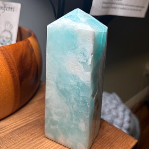 May include: A polished, tower-shaped crystal point featuring a blend of opaque white and translucent light blue mineral patterns. The stone has a hexagonal base and a pointed top, showcasing natural variations in color and texture throughout its surface.