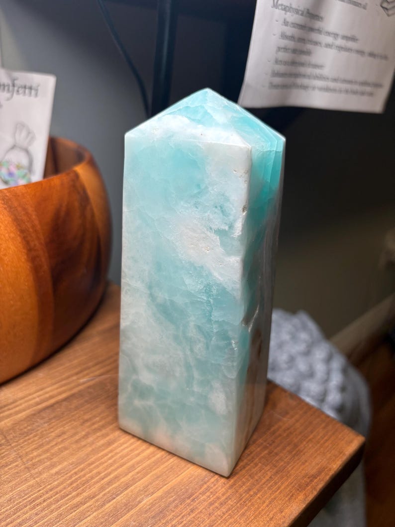 May include: A polished, tower-shaped crystal point featuring a blend of opaque white and translucent light blue mineral patterns. The stone has a hexagonal base and a pointed top, showcasing natural variations in color and texture throughout its surface.