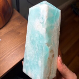 May include: A polished, hexagonal crystal tower featuring a vibrant turquoise and light blue color palette with opaque white mineral inclusions and subtle brown veining running vertically along one side of the smooth, translucent stone surface.