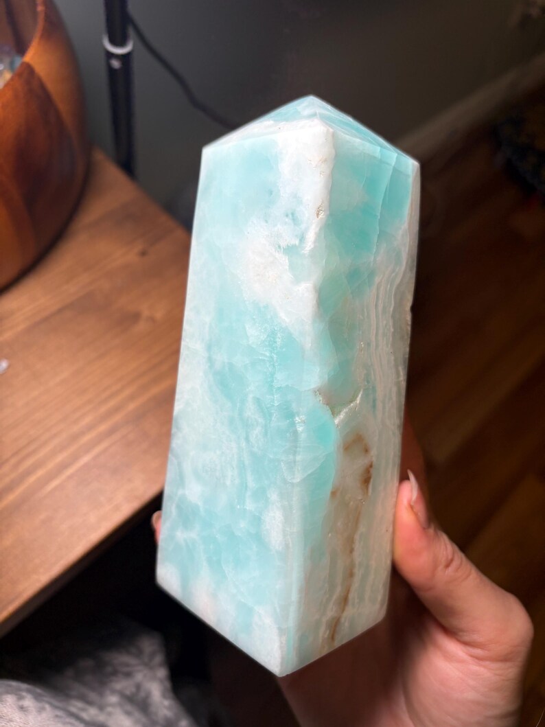 May include: A polished, hexagonal crystal tower featuring a vibrant turquoise and light blue color palette with opaque white mineral inclusions and subtle brown veining running vertically along one side of the smooth, translucent stone surface.