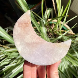 May include: A pale pink and white crescent moon shaped crystal. The crystal is smooth and polished. It is being held in a hand.