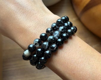 May include: A set of three stretch bracelets composed of polished, spherical beads in shades of black, charcoal, and dark gray. The beads feature natural inclusions and subtle marbling patterns, creating a varied texture across the dark surface.