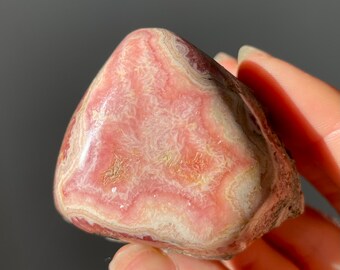 May include: A pink and white banded polished stone with a swirling pattern. The stone is smooth and has a natural, organic shape.