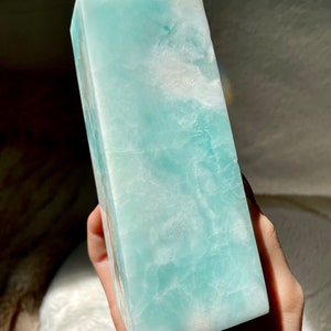 May include: A polished, tower-shaped crystal point featuring a smooth, translucent surface with a marbled pattern of light turquoise, pale blue, and opaque white mineral inclusions throughout the stone structure.