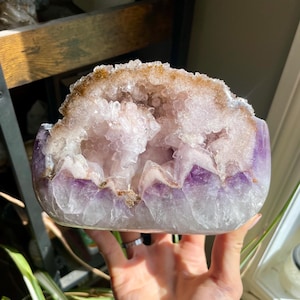 May include: A large amethyst geode with a polished exterior. The geode features a deep purple rim that fades into a light lavender interior filled with sparkling crystal formations. The top edge has a band of golden-brown druzy.