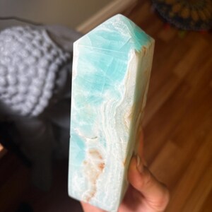 May include: A polished Caribbean calcite crystal tower featuring a hexagonal point. The stone displays a vibrant turquoise blue color with translucent layers and natural white and tan mineral inclusions running throughout the vertical structure.